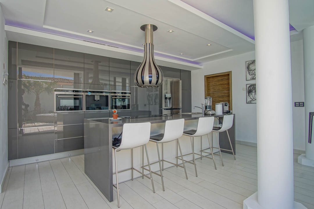 Cocina moderna con armarios grises de alto brillo, electrodomésticos integrados y una isla central con barra de desayuno para cuatro. Iluminación LED y vistas reflejadas al exterior.