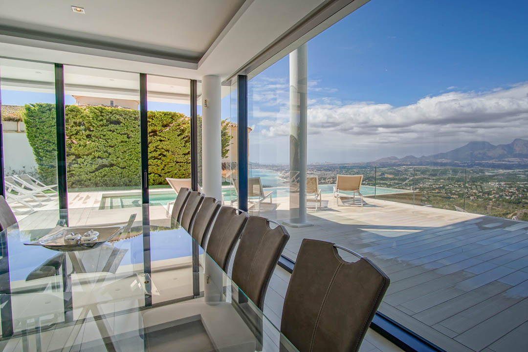 Comedor interior elegante con mesa de cristal y sillas, conectado a una amplia terraza con piscina infinita y vistas panorámicas al mar y la montaña.