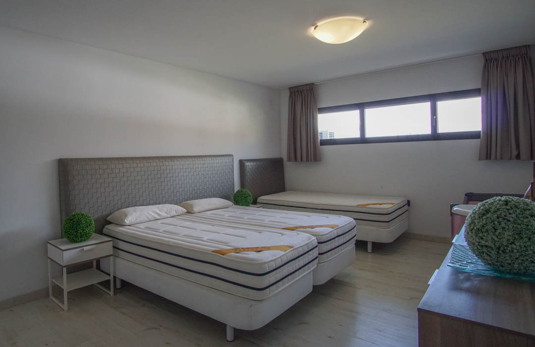 Dormitorio con dos camas individuales unidas, cabeceros grises texturizados, una tercera cama individual al fondo, suelo de madera clara y ventanas horizontales.