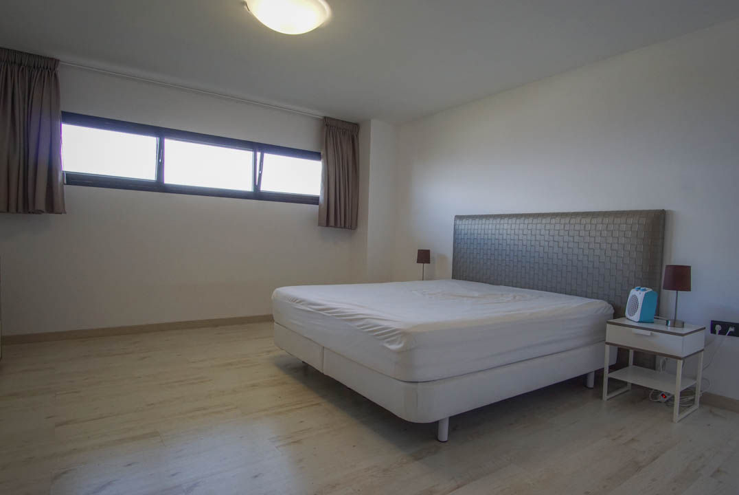 Dormitorio con paredes blancas y suelo de madera clara. Incluye una cama con cabecero texturizado, mesita de noche con lámpara y una ventana horizontal con cortinas.