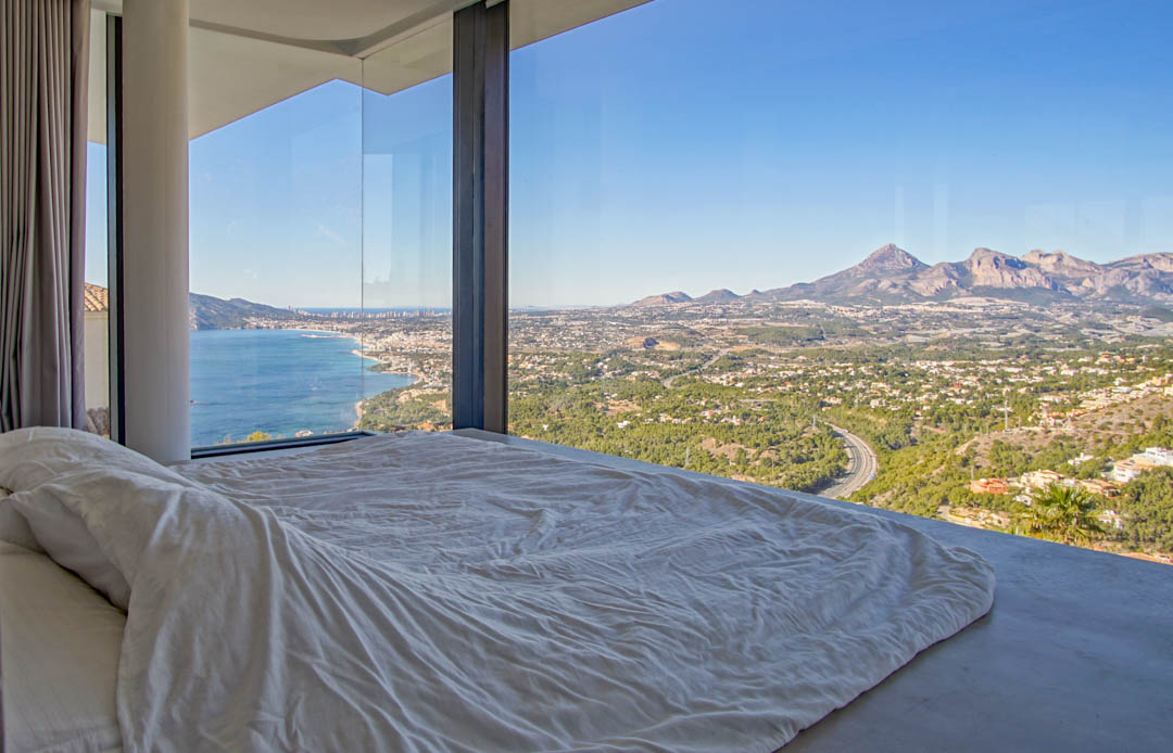 Dormitorio con ventanales de suelo a techo, ofreciendo vistas panorámicas al mar Mediterráneo, una ciudad costera y montañas lejanas. Una cama baja con ropa blanca mira hacia el horizonte.