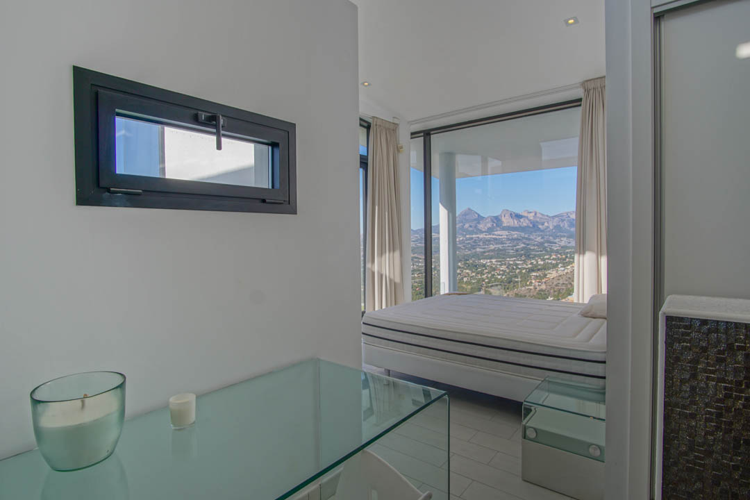 Dormitorio luminoso con cama frente a ventanales de suelo a techo, revelando vistas panorámicas a la montaña y el valle. Se aprecia una consola de cristal y una ventana horizontal con marco oscuro.