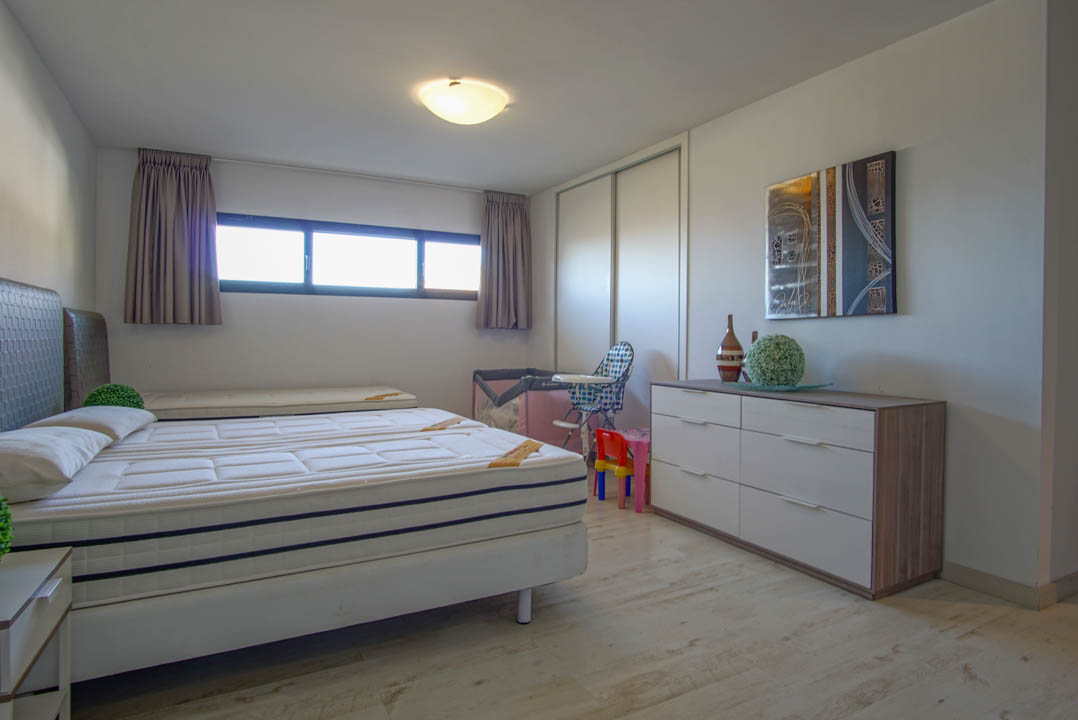 Dormitorio luminoso con una cama de dos colchones, suelo de madera clara y una ventana horizontal alargada. Incluye un armario blanco de puertas correderas y una cómoda moderna con arte abstracto.