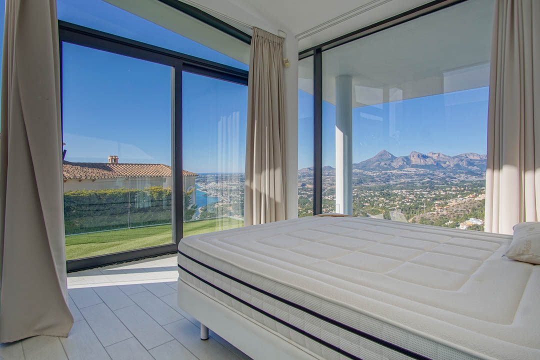 Dormitorio moderno con cama grande y amplios ventanales. Ofrece vistas panorámicas al mar distante, un césped verde y montañas escarpadas bajo un cielo despejado.