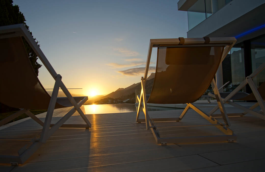 Dos tumbonas modernas sobre tarima clara frente a una piscina infinita que refleja el atardecer dorado sobre las montañas.
