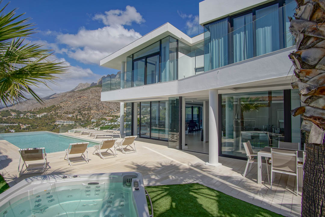 El exterior de la villa moderna blanca presenta una piscina infinita, jacuzzi independiente y una amplia terraza con tumbonas, enmarcado por vistas a la montaña bajo un cielo despejado.