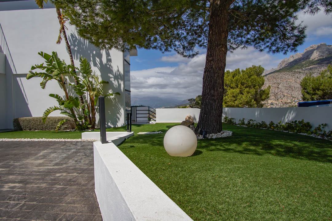 El exterior de la villa presenta una entrada pavimentada, césped artificial con un pino maduro, una moderna luz esférica y vistas a la montaña.