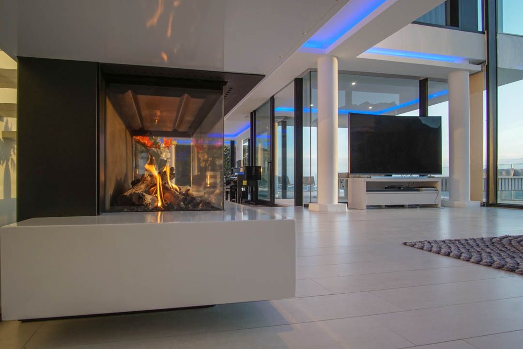 Espacio de salón moderno con chimenea de gas acristalada y base blanca. Grandes ventanales y luz ambiental azul complementan el diseño.