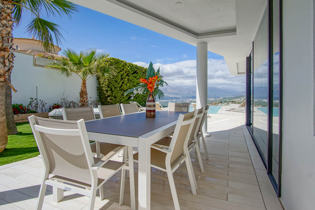 Esta terraza comedor exterior cubierta presenta una mesa blanca moderna con ocho sillas beige sobre baldosas claras. Ofrece amplias vistas a las montañas y al mar, junto a la piscina infinita.