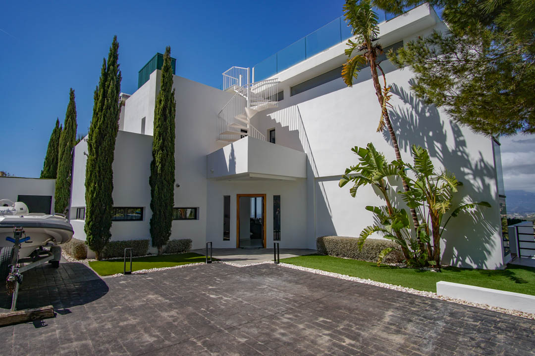 Exterior de la villa moderna con entrada pavimentada, cipreses altos y una escalera de caracol blanca que asciende a la terraza superior con barandilla de cristal.