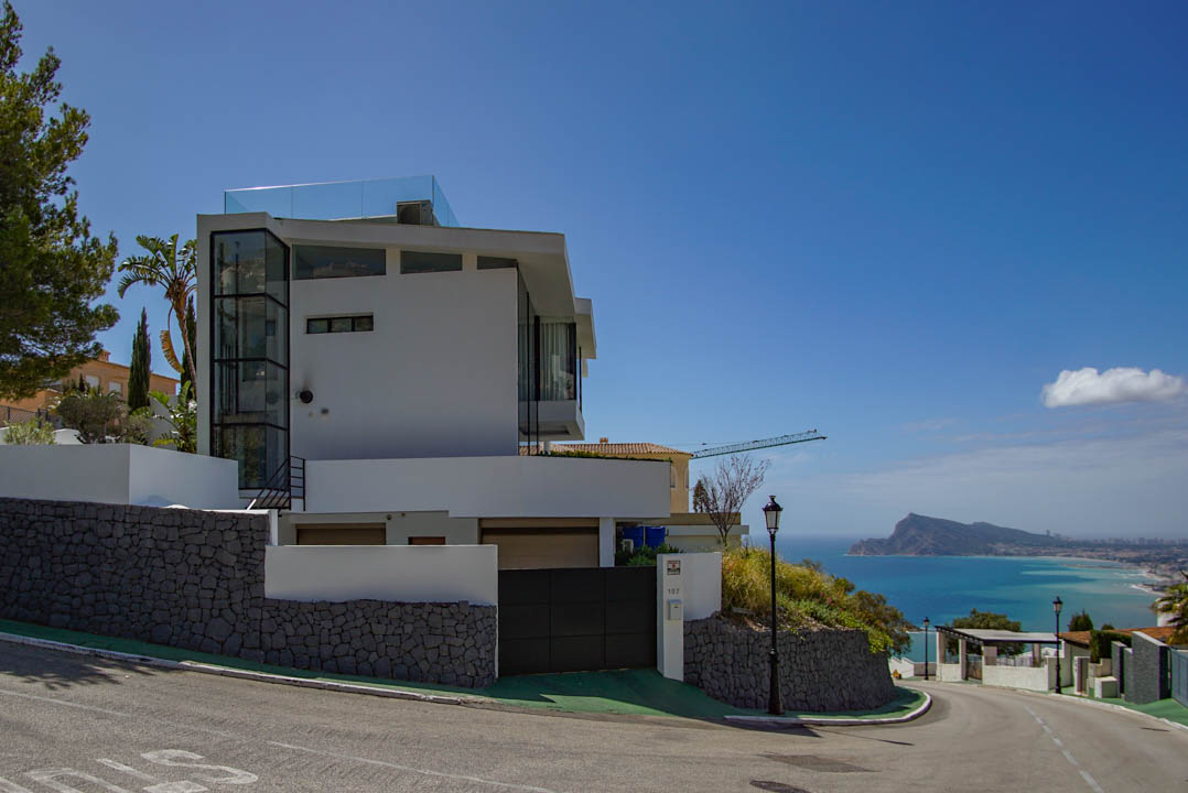 Exterior de villa moderna blanca con ascensor de cristal visible y barandilla de terraza en la azotea. Muros de piedra y portón automático complementan las vistas panorámicas al mar.