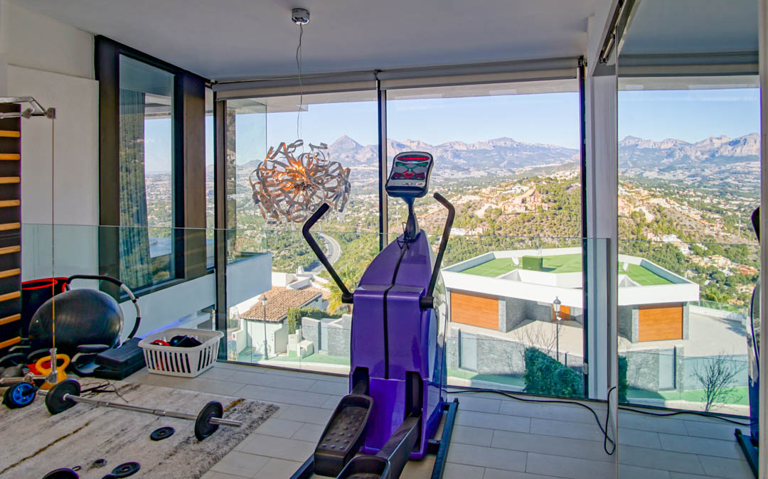 Gimnasio con ventanales de suelo a techo, ofreciendo vistas panorámicas a las montañas y al valle. Equipado con elíptica y pesas, el espacio incluye una barandilla de cristal y un diseño moderno.