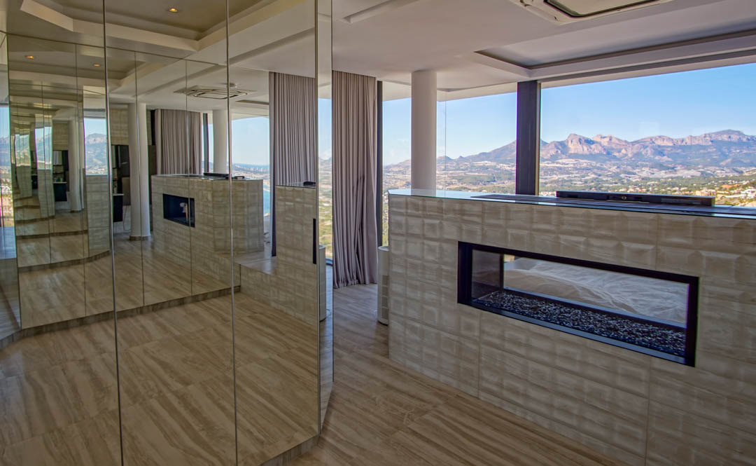 Interior moderno con chimenea de gas transparente y base de guijarros, integrada en una pared texturizada. Paneles espejados reflejan la estancia y las amplias vistas a la montaña y al mar.