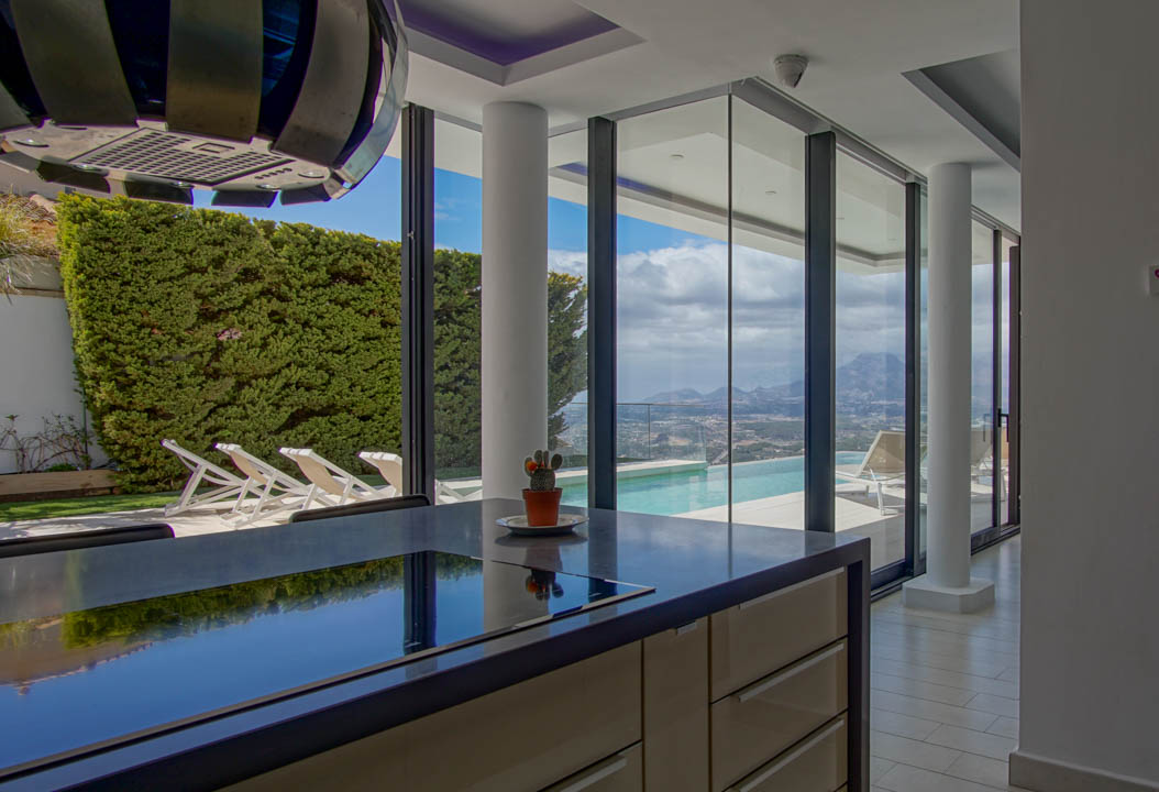 Isla de cocina moderna con encimera oscura y placa integrada, frente a grandes ventanales con vistas a la piscina infinita, el valle y las montañas.