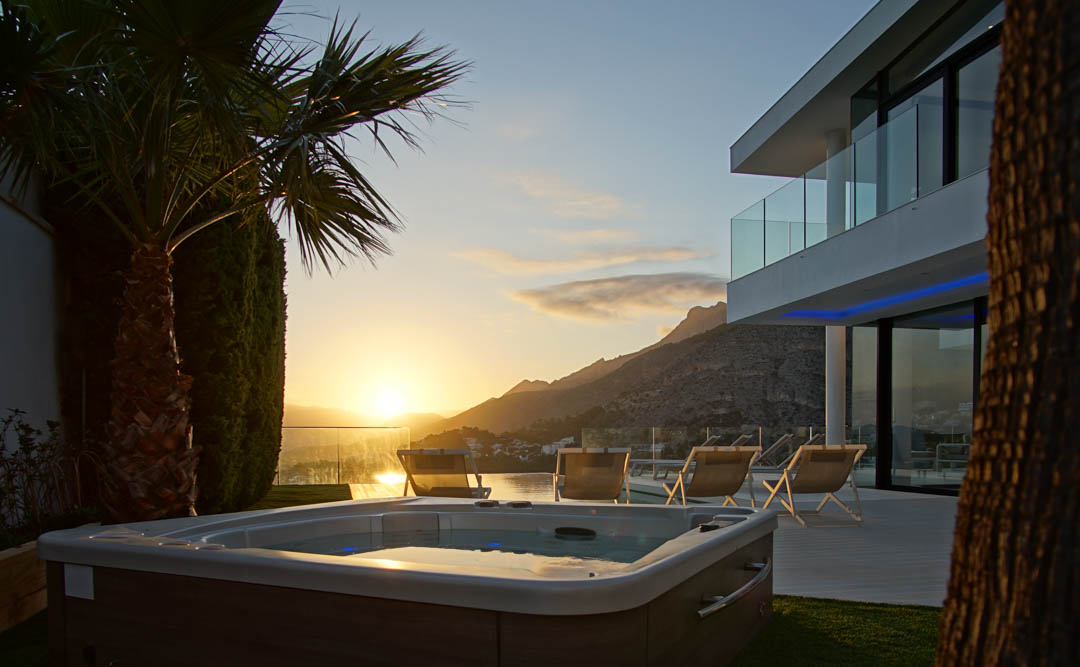 Jacuzzi exterior con acabado de madera, rodeado de césped, frente a una villa moderna con balcones de cristal. Vistas a la montaña y al mar al atardecer.