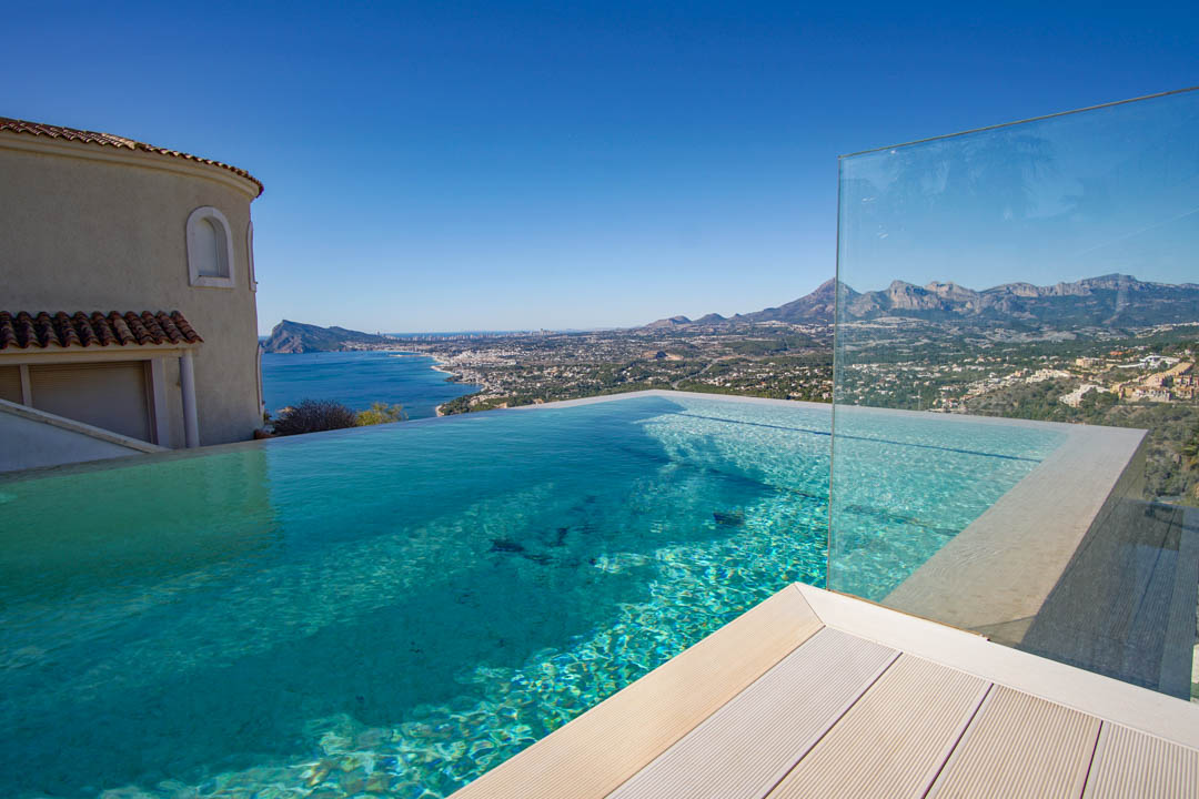 La piscina infinita de agua cristalina y cubierta clara, con barandilla de cristal, ofrece vistas panorámicas al mar, la costa y las montañas bajo un cielo despejado.