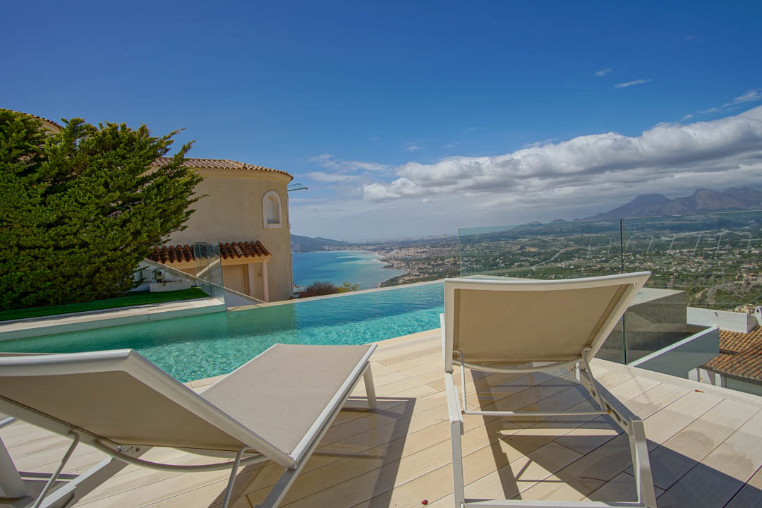 La terraza exterior presenta una piscina infinita con vistas a la costa y montañas lejanas. Dos tumbonas sobre tarima clara, enmarcadas por barandilla de cristal y fachada de villa tradicional.