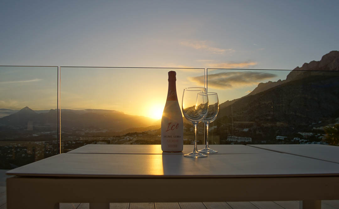 Mesa exterior en terraza con barandilla de cristal, presentando una botella de cava y dos copas. Atardecer sobre montañas y valle, con el sol poniente reflejándose.