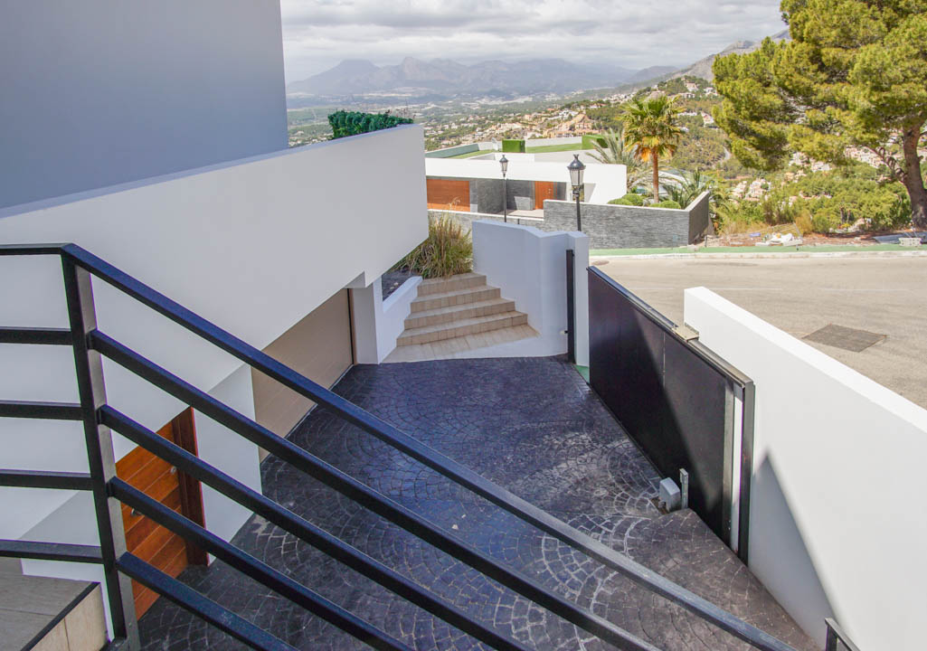 Moderna escalera exterior con barandales negros desciende a un camino de entrada texturizado en gris oscuro, con puerta automática, acceso a garaje y vistas panorámicas a la montaña.