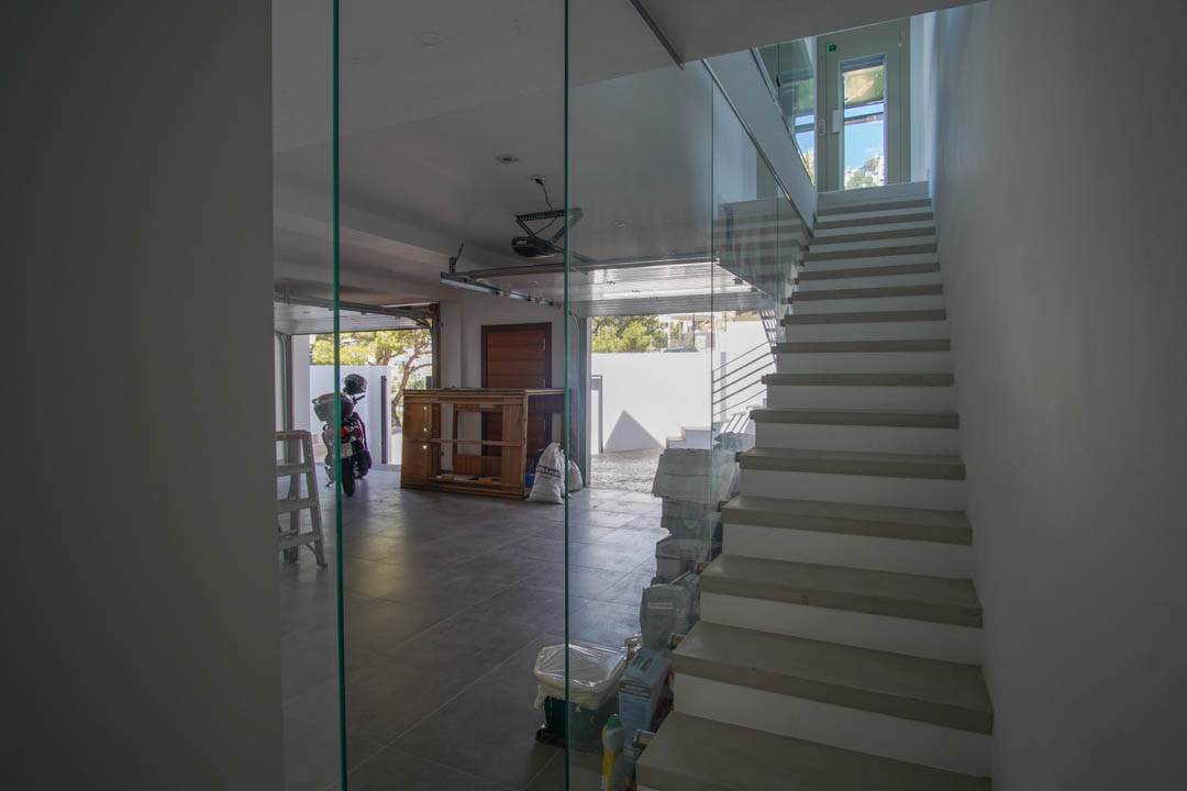 Moderna escalera interior con barandilla de cristal y peldaños claros, adyacente a un garaje con suelo de baldosas y puerta abierta con luz natural.