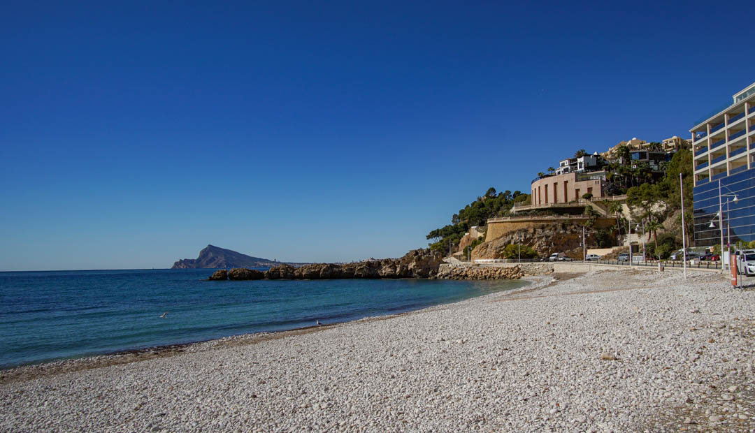 Playa de guijarros con aguas turquesas cristalinas y costa rocosa. Edificios modernos de fachadas reflectantes y una estructura redondeada bordean la ladera, bajo un cielo azul.