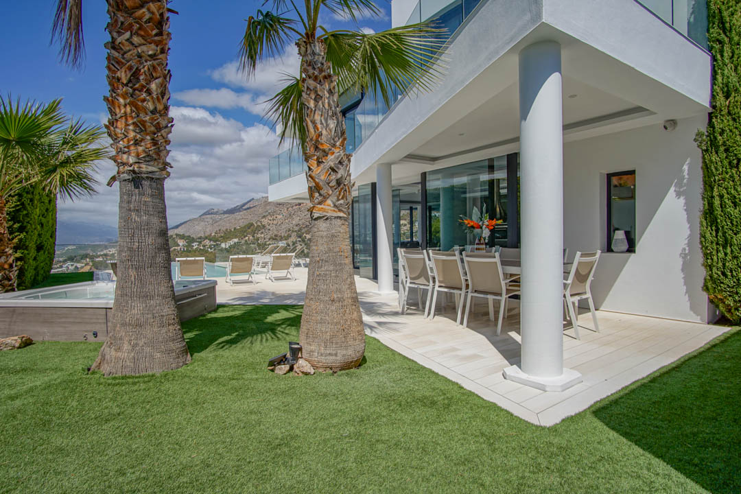 Terraza comedor exterior cubierta con mobiliario moderno, junto a un césped artificial y palmeras. La zona de jacuzzi y piscina ofrece vistas a la montaña y al mar.