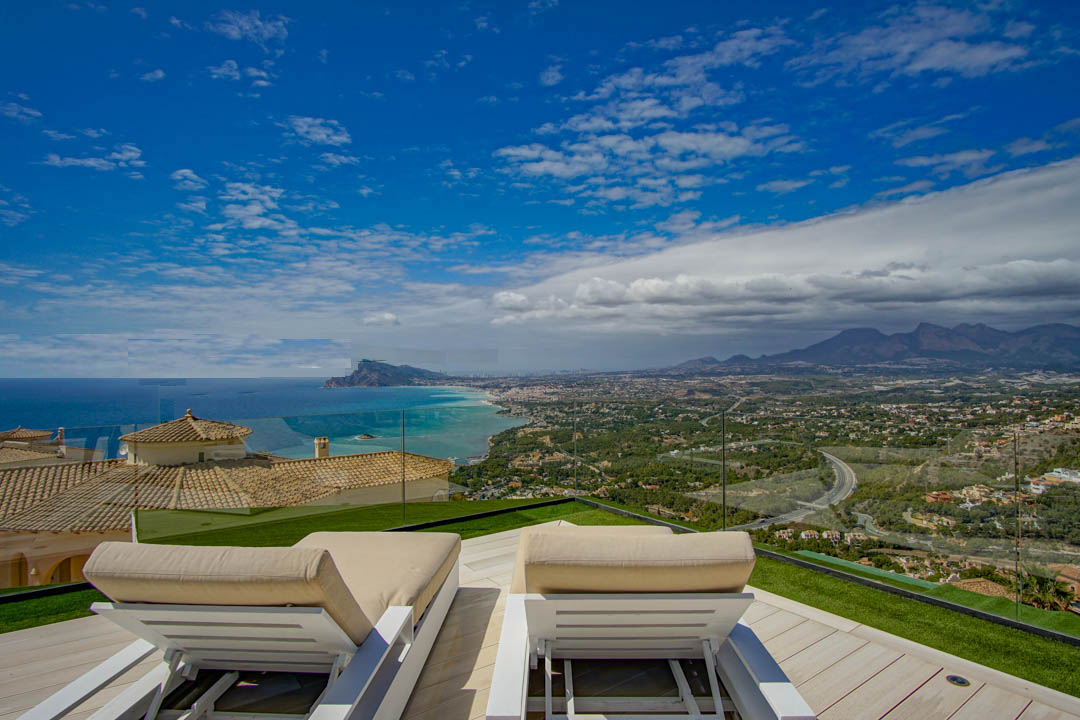 Terraza en la azotea con dos tumbonas blancas sobre tarima de madera clara y césped artificial, con vistas panorámicas al Mediterráneo, la costa de Altea y las montañas lejanas.