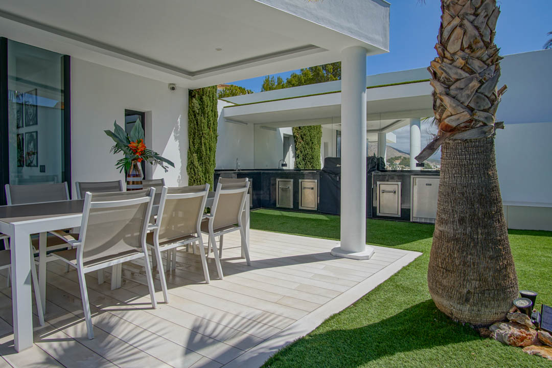 Terraza exterior con comedor al aire libre sobre suelo de lamas claras y césped artificial. Incluye una cocina exterior cubierta con barbacoa y electrodomésticos de acero inoxidable, junto a una palmera.