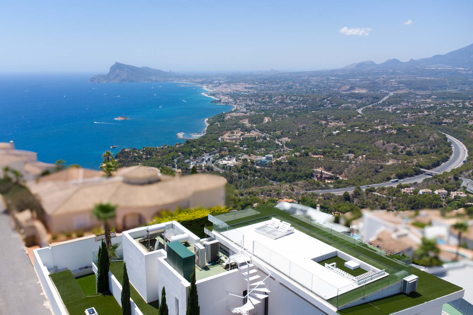 Vista aérea de la terraza en la azotea de la villa con escalera de caracol blanca, césped artificial, barandillas de cristal y vistas panorámicas al mar Mediterráneo y la costa.