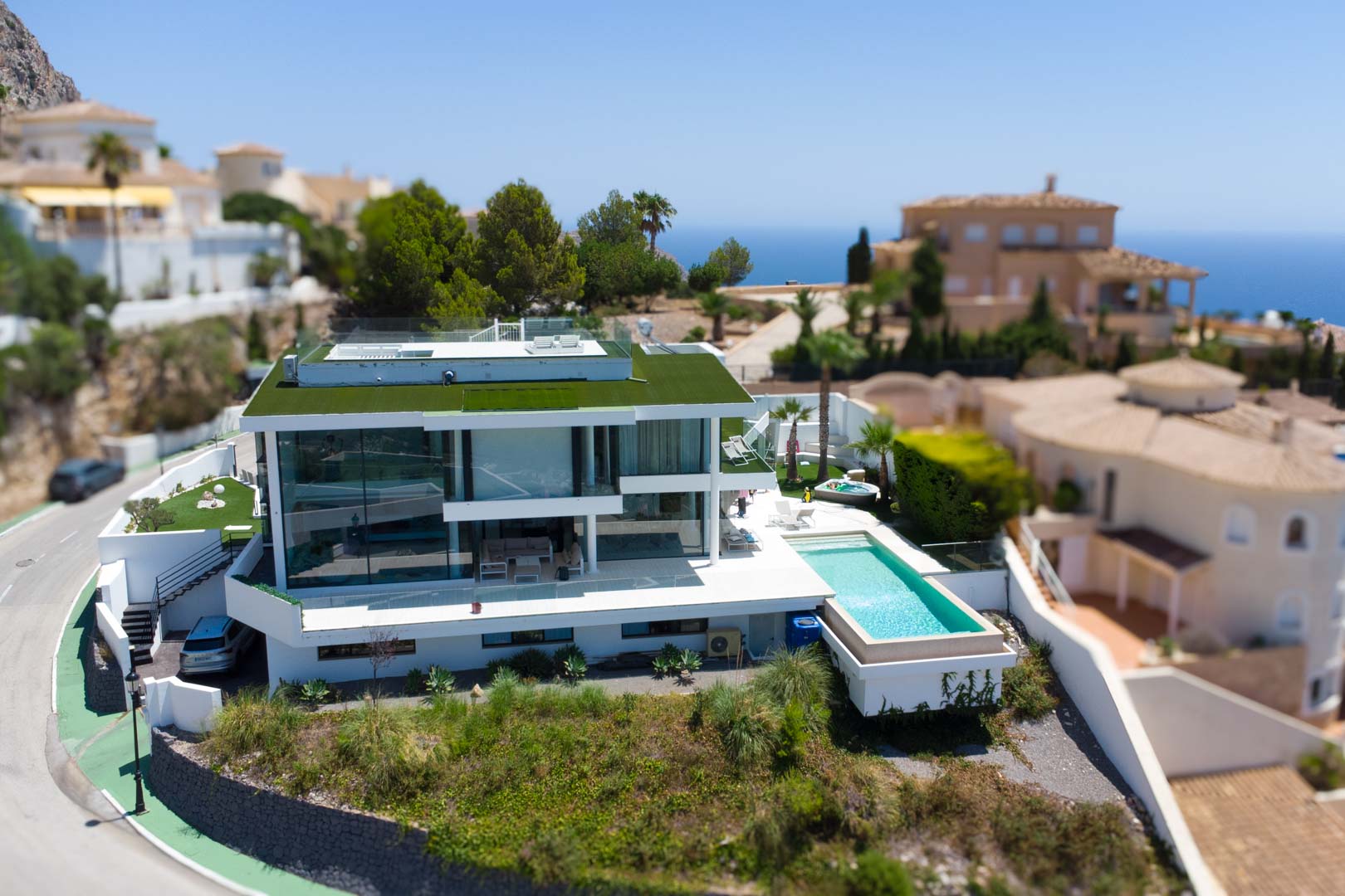 Vista aérea de la villa moderna de diseño blanco con techo verde, piscina infinita y jacuzzi separado, en un entorno con vistas al mar.