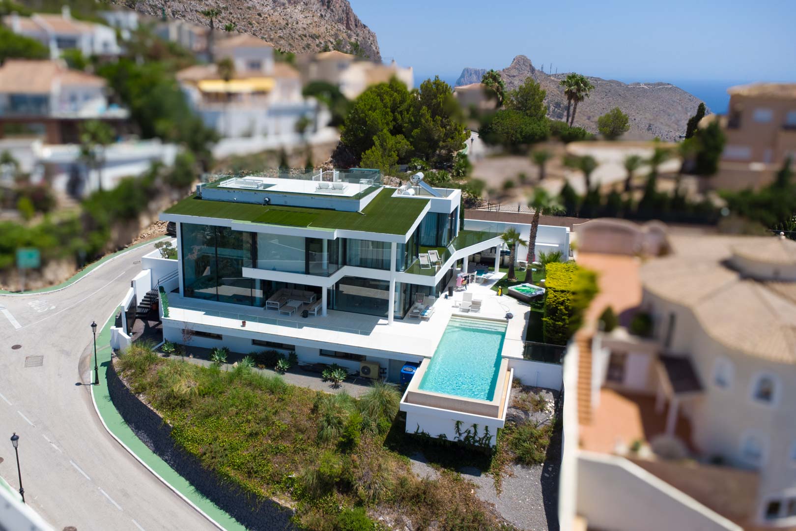 Vista aérea de una villa blanca moderna con piscina infinita, amplias fachadas de cristal, techo verde y jacuzzi, ofreciendo vistas a la montaña y al mar.