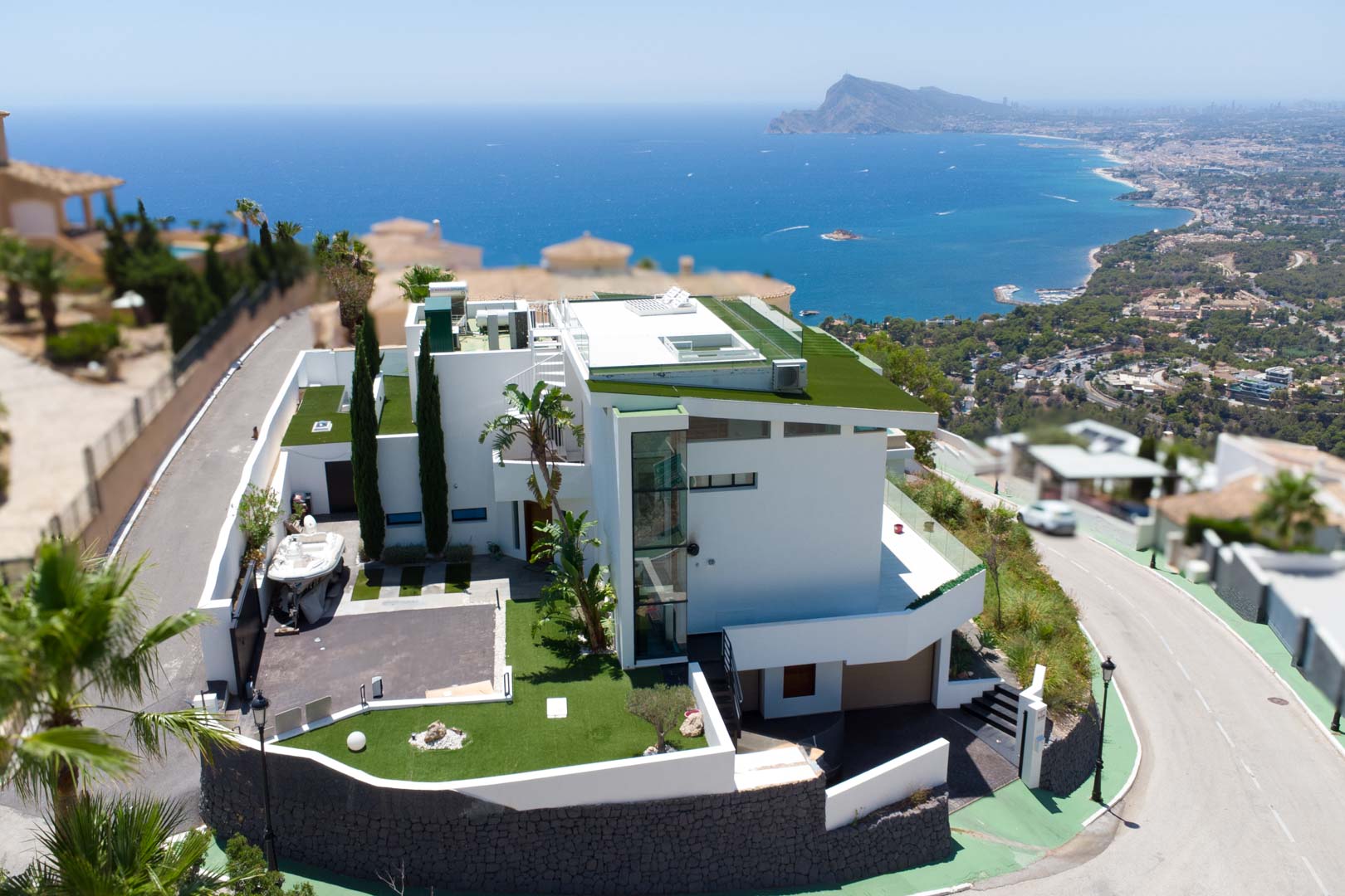 Vista aérea de una villa moderna blanca con arquitectura de varios niveles, techos planos verdes y ascensor exterior de cristal. La propiedad incluye entrada privada y ofrece vistas panorámicas al mar y la costa.