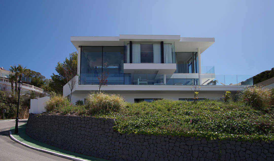 Vista exterior de villa moderna de tres niveles con amplias fachadas de cristal y barandillas de vidrio, elevada sobre un muro de piedra y vegetación junto a una carretera curva.