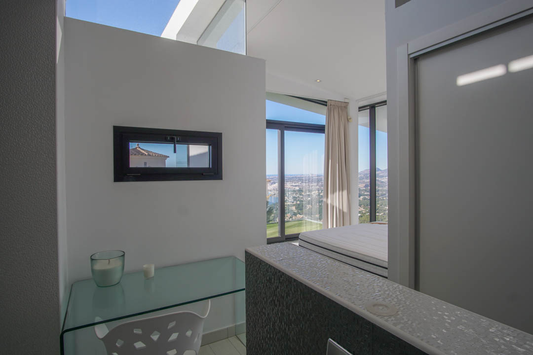 Zona de estudio con escritorio de cristal y ventana horizontal, ofreciendo vistas panorámicas al mar y la ciudad desde el dormitorio adyacente.