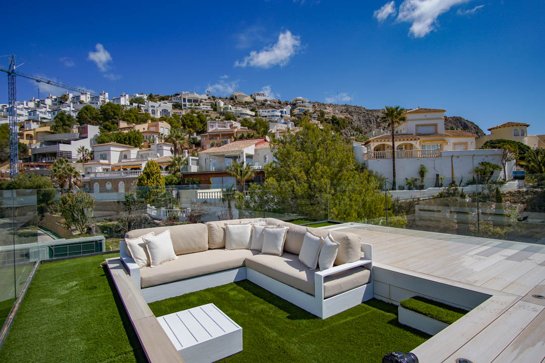 Zona de salón en la azotea con sofá en forma de L sobre césped artificial, mesa de centro blanca y barandillas de cristal. Vistas despejadas a la ladera escalonada con viviendas residenciales.