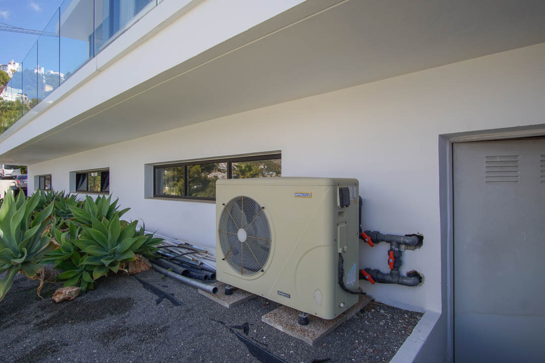 Zona técnica exterior de la villa con bomba de calor inverter AstralPool, ventanas horizontales y puerta de servicio. Agaves adornan la moderna fachada blanca.