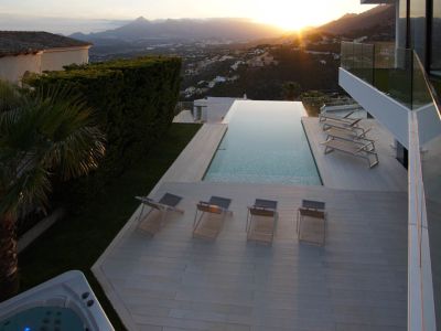 Amplia piscina infinita con tarima clara, varias tumbonas y jacuzzi separado, enmarcada por un paisaje montañoso con puesta de sol panorámica.