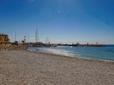 Amplia playa de guijarros se extiende hacia un puerto deportivo con yates, flanqueada por edificios costeros tradicionales bajo un cielo azul claro.