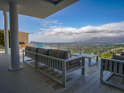 Amplia terraza cubierta con tarima clara y mobiliario lounge blanco, enmarcando una vista panorámica al mar, la costa y las montañas a través de una barandilla de cristal.