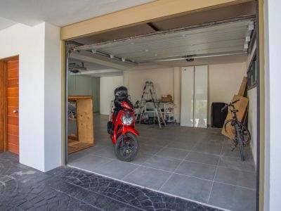 Amplio garaje con suelo de baldosas grises, puerta seccional parcialmente abierta y una puerta de entrada de madera a la izquierda. En el interior se ven una moto roja y una bicicleta.
