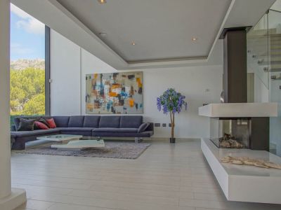 Amplio salón con sofá gris en forma de L, mesas de centro modernas y chimenea de gas. Grandes ventanales ofrecen vistas a la montaña y árboles, junto a una escalera de cristal.