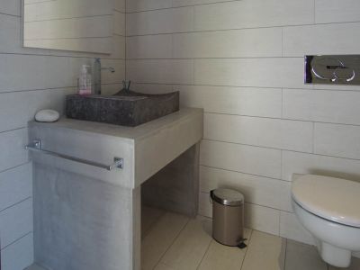 Aseo moderno con lavabo de piedra sobre encimera de hormigón y azulejos claros. Inodoro suspendido con pulsador cromado.