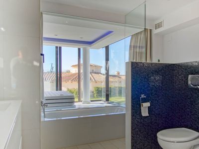 Baño moderno con bañera integrada y ventanales de suelo a techo que ofrecen vistas a un tejado de tejas y jardín. Dispone de iluminación LED azul y una pared divisoria con patrón oscuro.