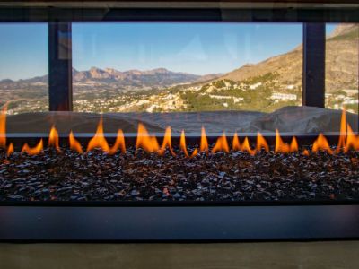 Chimenea de gas moderna con llamas sobre cristales oscuros, enmarcando una vista panorámica a montañas y un valle.