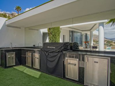 Cocina exterior en forma de L con encimeras de granito negro, fregaderos dobles, barbacoa cubierta y electrodomésticos de acero inoxidable, con vistas reflejadas a la piscina.
