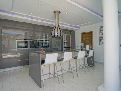 Cocina moderna con armarios grises de alto brillo, electrodomésticos integrados y una isla central con barra de desayuno para cuatro. Iluminación LED y vistas reflejadas al exterior.