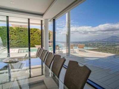 Comedor interior elegante con mesa de cristal y sillas, conectado a una amplia terraza con piscina infinita y vistas panorámicas al mar y la montaña.