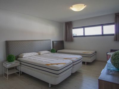 Dormitorio con dos camas individuales unidas, cabeceros grises texturizados, una tercera cama individual al fondo, suelo de madera clara y ventanas horizontales.