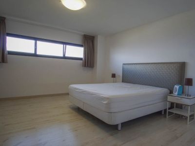 Dormitorio con paredes blancas y suelo de madera clara. Incluye una cama con cabecero texturizado, mesita de noche con lámpara y una ventana horizontal con cortinas.