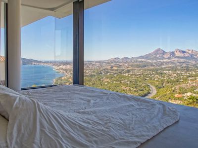 Dormitorio con ventanales de suelo a techo, ofreciendo vistas panorámicas al mar Mediterráneo, una ciudad costera y montañas lejanas. Una cama baja con ropa blanca mira hacia el horizonte.